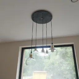 Nowoczesna lampa wisząca z pięcioma szklanymi kloszami o różnych kształtach, zamontowana na suficie w jasnym wnętrzu z widokiem na zieleń za oknem.