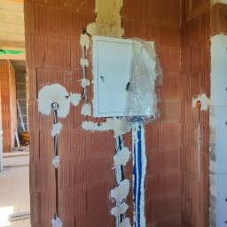 Paweł Bartoniek Twój Elektryk Rydułtowy - Rozprowadzona instalacja elektryczna w ścianie z cegły, z białą skrzynką zabezpieczeń, niebieskie rury kablowe, całość zabezpieczona pianką montażową, w trakcie prac wykończeniowych.