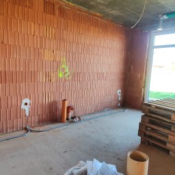 Paweł Bartoniek Twój Elektryk Rydułtowy - Surowy stan budynku z cegły: widoczne instalacje elektryczne w ścianach i rury kanalizacyjne, okno z paletami, betonowa podłoga i sufit. Oznakowania na ścianie.