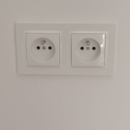 Paweł Bartoniek Twój Elektryk Rydułtowy - Białe, podwójne gniazdko elektryczne typu europejskiego, zamontowane na gładkiej, białej ścianie. Widoczne śruby mocujące i otwory na bolce uziemiające.