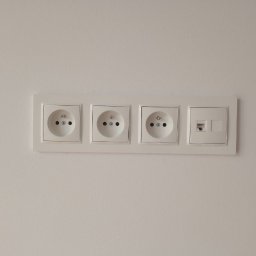 Paweł Bartoniek Twój Elektryk Rydułtowy - Biały panel z trzema gniazdkami elektrycznymi typu E i gniazdem sieciowym RJ45, zamontowany na jasnej ścianie. Minimalistyczny design, symetryczny układ.