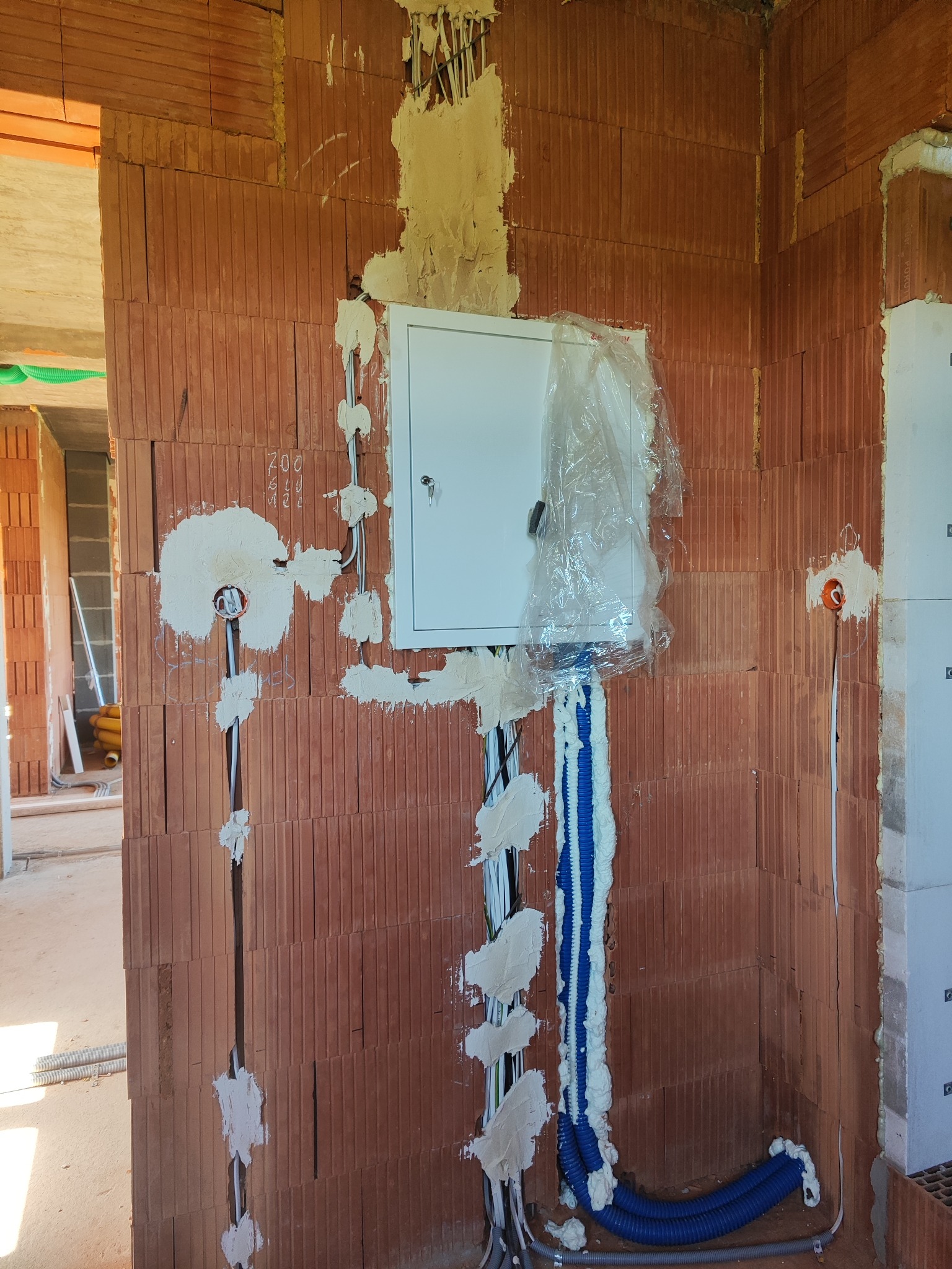Rozprowadzona instalacja elektryczna w ścianie z cegły, z białą skrzynką zabezpieczeń, niebieskie rury kablowe, całość zabezpieczona pianką montażową, w trakcie prac wykończeniowych.