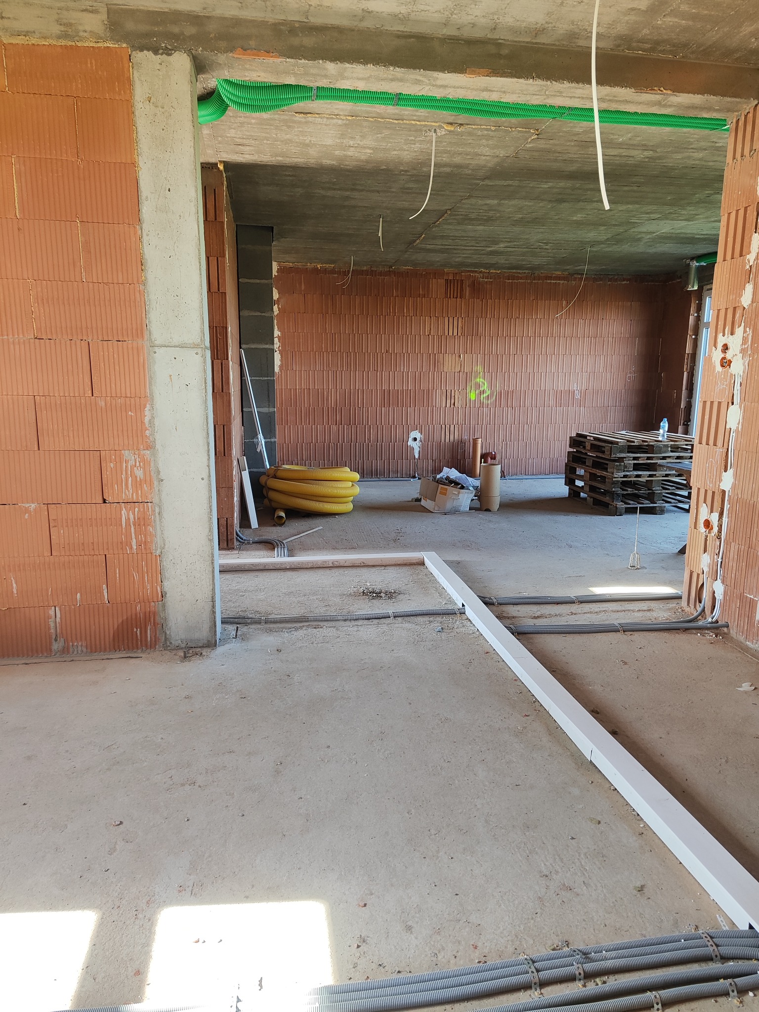 Wnętrze budynku w trakcie budowy z widocznymi przewodami instalacji elektrycznej w ścianach i pod sufitem, oraz rurami na podłodze. Ceglane ściany i surowy beton.