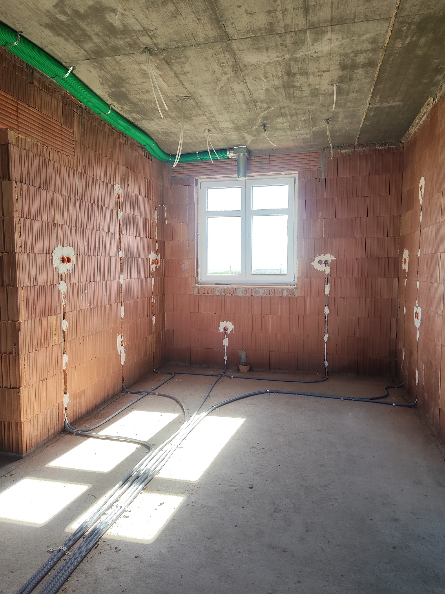 Surowy stan: instalacja elektryczna w nowym budynku z cegły, widoczne kable w ścianach i podłodze, rury wentylacyjne pod sufitem, okno w tle.