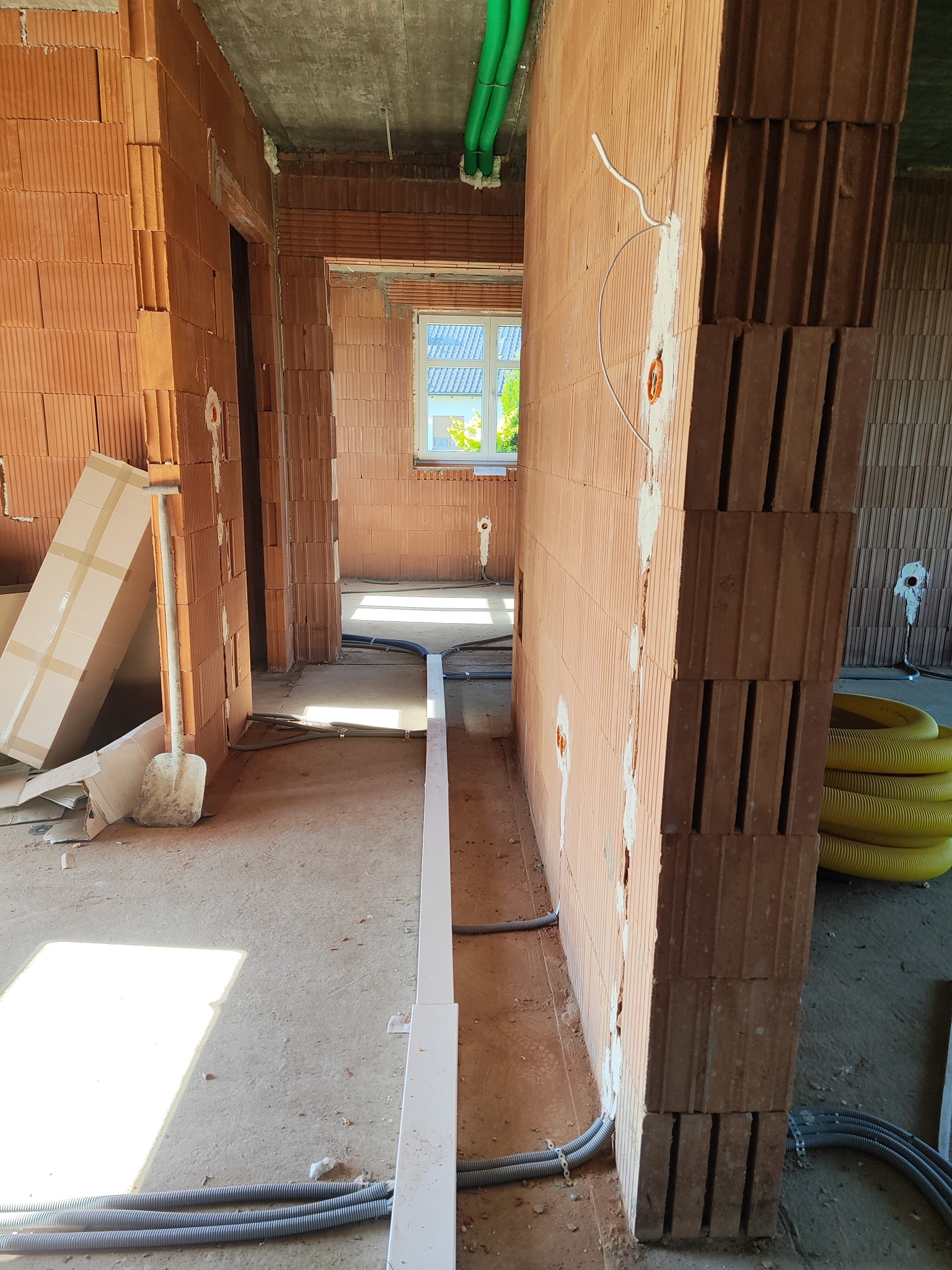 Wnętrze w trakcie budowy z instalacją elektryczną w ścianach z cegły. Widoczne kable i rury w korytarzu, okno w tle i narzędzia budowlane.