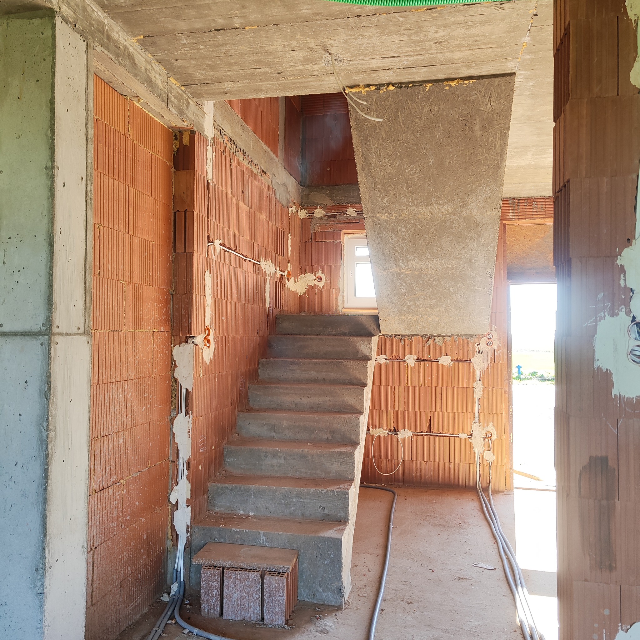 Surowy stan domu w budowie: widoczne ceglane ściany z bruzdami na przewody, betonowe schody i wystające kable. W tle okno i otwarte drzwi.
