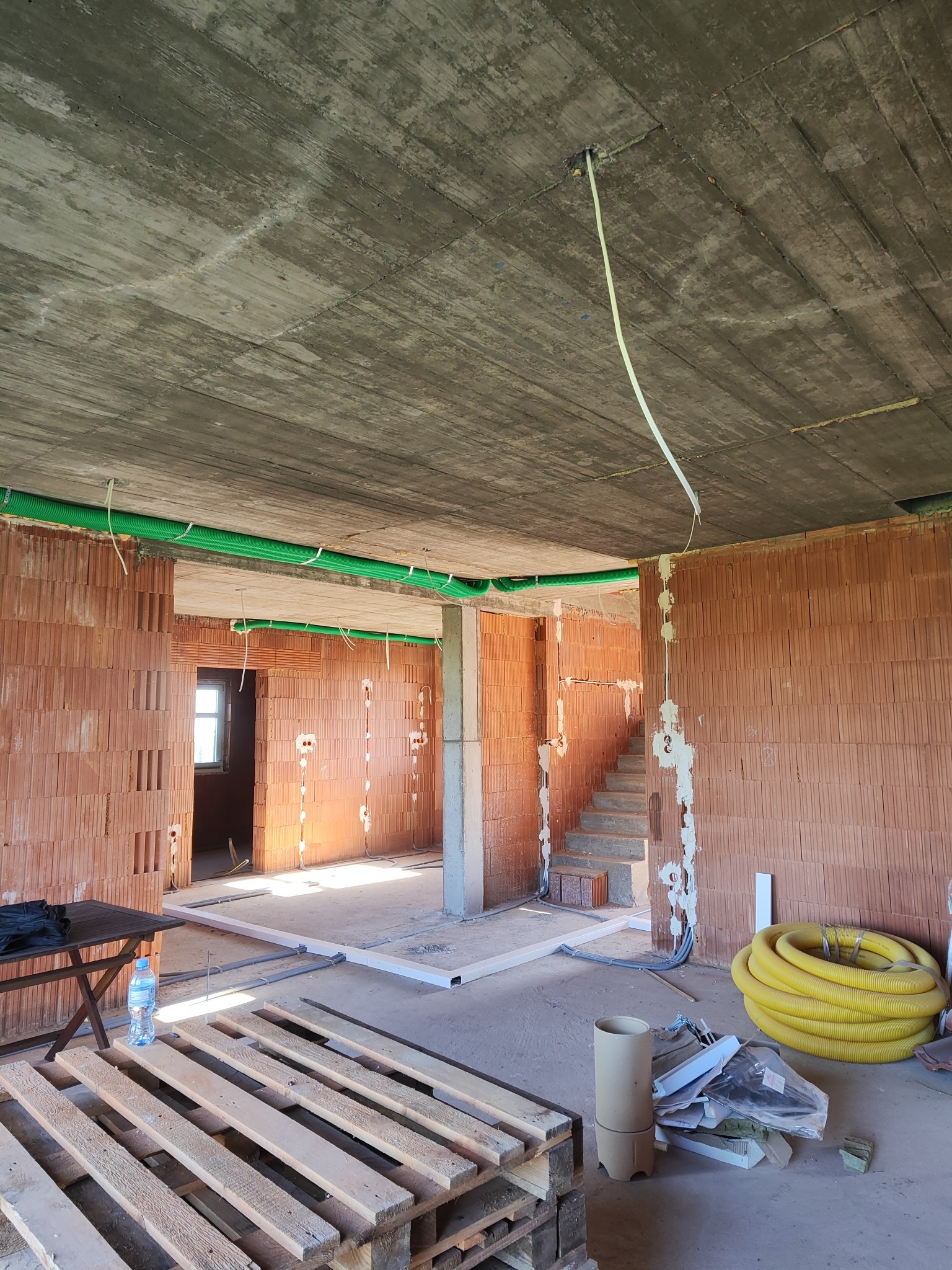 Surowy stan wnętrza domu w trakcie instalacji elektrycznej: zielone rury na ścianach, przewody wychodzące z sufitu, puszki elektryczne w murze z cegły.