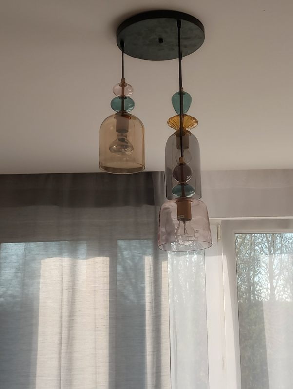 Dwa wiszące, szklane żyrandole z kolorowymi elementami dekoracyjnymi, oświetlające wnętrze z oknem w tle. Jeden żyrandol ma klosz w odcieniach brązu, a drugi szarości.