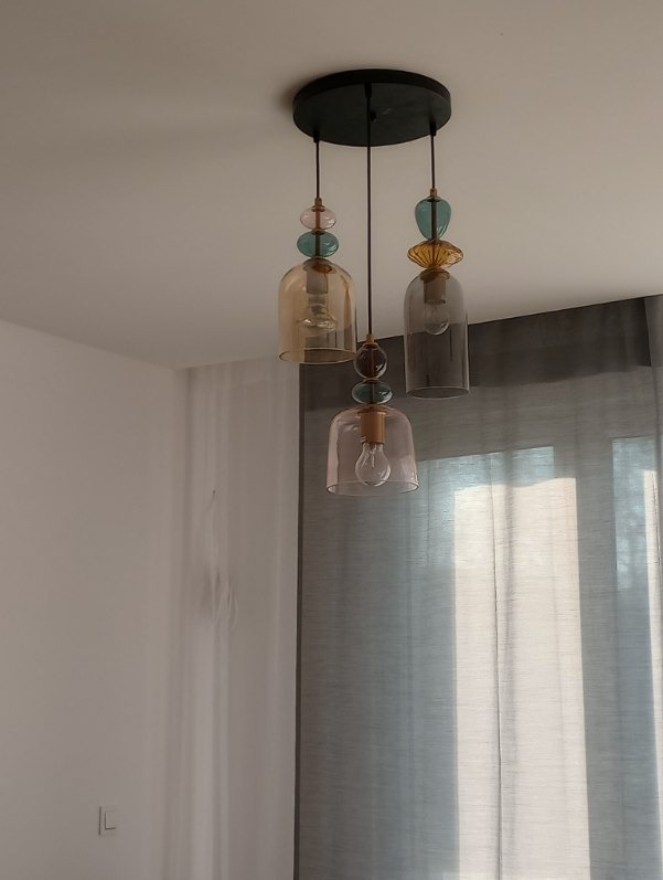 Trzy lampy wiszące z kloszami w pastelowych odcieniach, zawieszone na czarnej, okrągłej podsufitce. Klosze w kształcie dzwonów, z dekoracyjnymi elementami na górze. Minimalistyczne wnętrze.