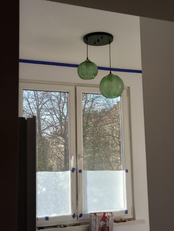 Dwa zielone, szklane klosze lamp wiszących nad oknem z częściowo zasłoniętą szybą, w pokoju z niebieskim paskiem dekoracyjnym pod sufitem.