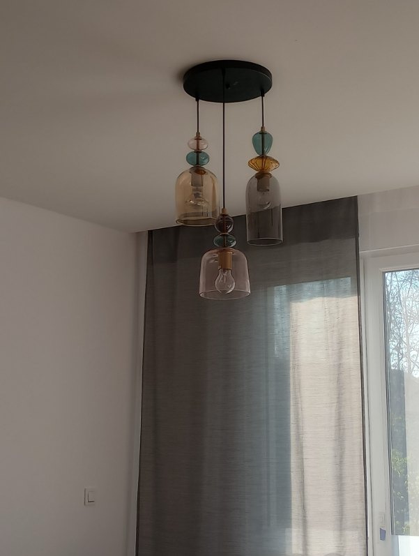 Trzyczęściowa lampa wisząca z kloszami w odcieniach bursztynu, różu i szarości, ozdobiona szklanymi kulkami, zawieszona na tle zasłony i okna.