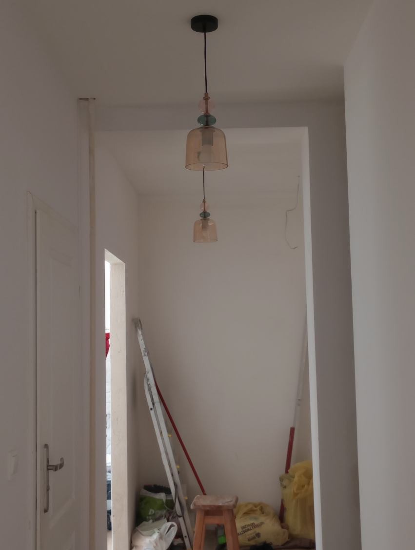 Dwupoziomowa lampa wisząca w odcieniach beżu i różu, zamontowana w jasnym wnętrzu. W tle widoczne drabina i inne sprzęty remontowe.