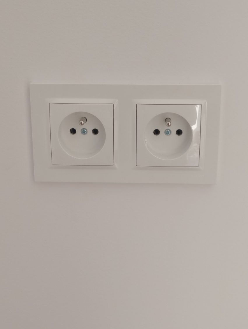 Białe, podwójne gniazdko elektryczne typu europejskiego, zamontowane na gładkiej, białej ścianie. Widoczne śruby mocujące i otwory na bolce uziemiające.