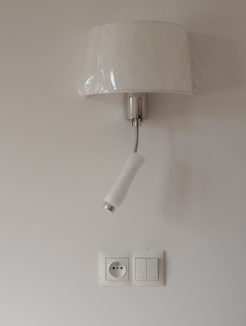 Ścienna lampka z abażurem w folii ochronnej, zainstalowana nad gniazdkiem i włącznikiem na białej ścianie. Lampa na elastycznym ramieniu.