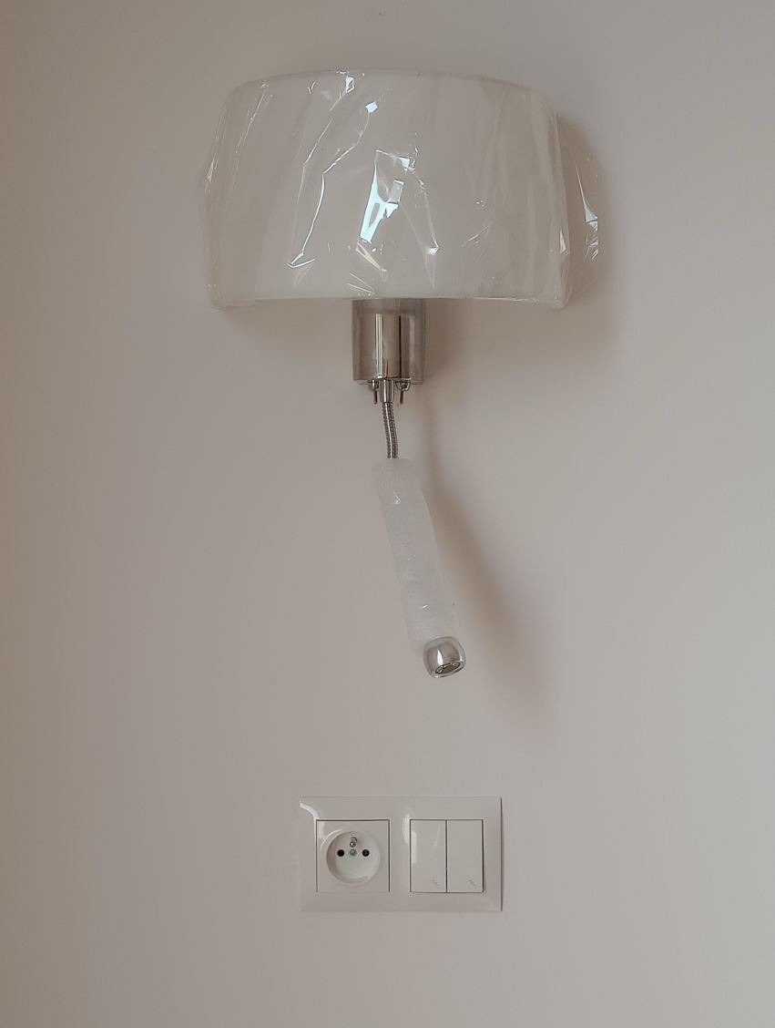 Kinkiet z elastycznym ramieniem, zamontowany na ścianie nad gniazdkiem i włącznikiem światła. Lampa zabezpieczona folią ochronną.