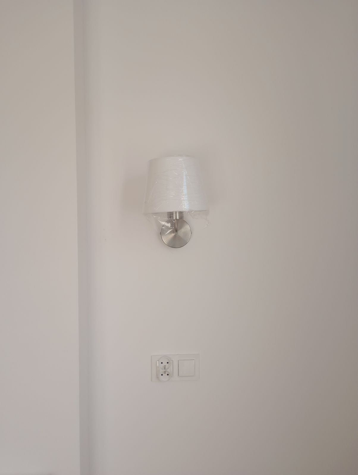 Ścienny kinkiet z abażurem, w folii ochronnej, zamontowany na białej ścianie. Poniżej gniazdko i włącznik. Widoczny fragment narożnika ściany.