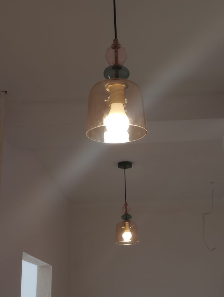 Dwa szklane, wiszące żyrandole z ozdobnymi elementami (różowa i zielona kula) i kloszem w kolorze bursztynowym, oświetlające wnętrze. Jeden żyrandol jest większy, drugi mniejszy.