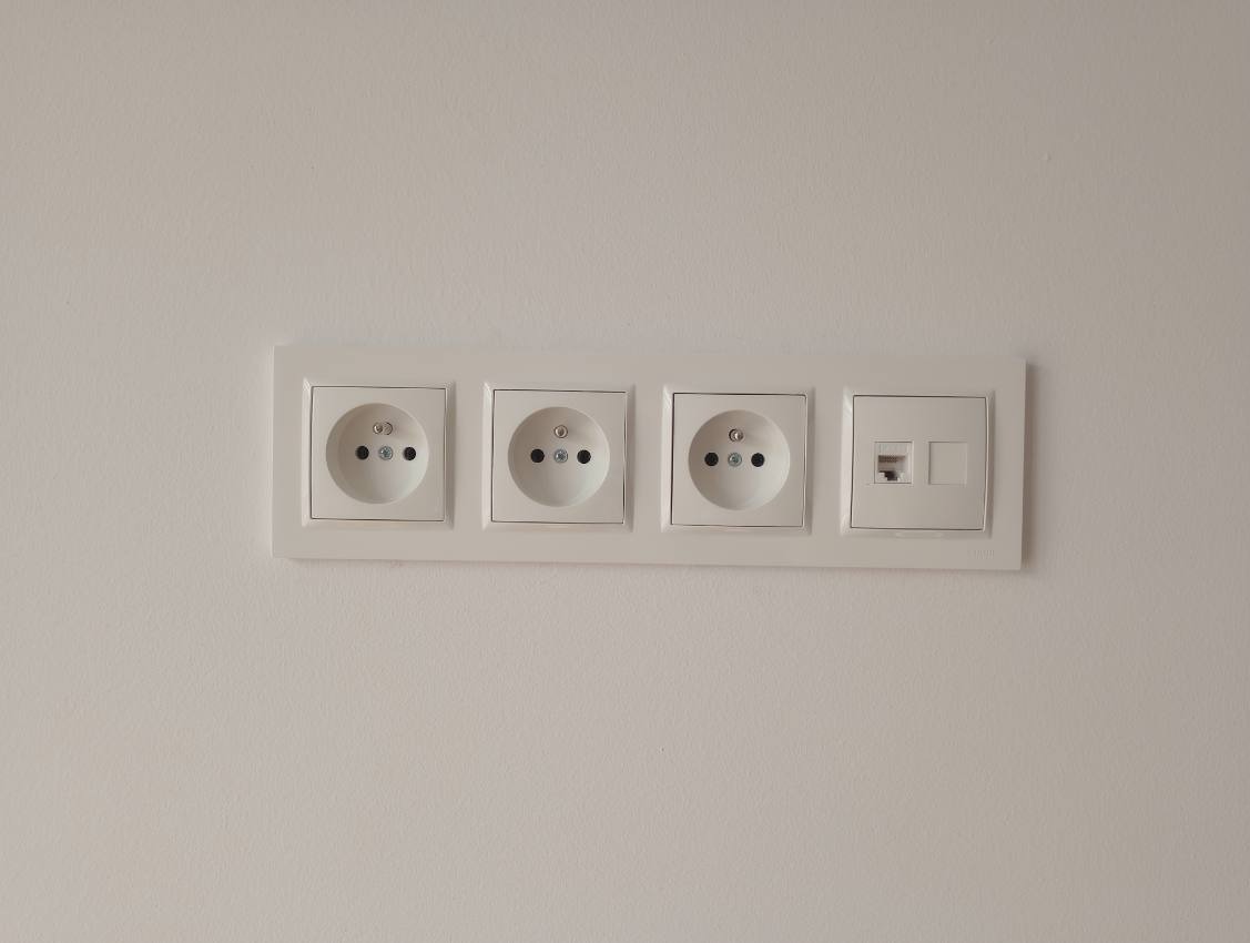Biały panel z trzema gniazdkami elektrycznymi typu E i gniazdem sieciowym RJ45, zamontowany na jasnej ścianie. Minimalistyczny design, symetryczny układ.