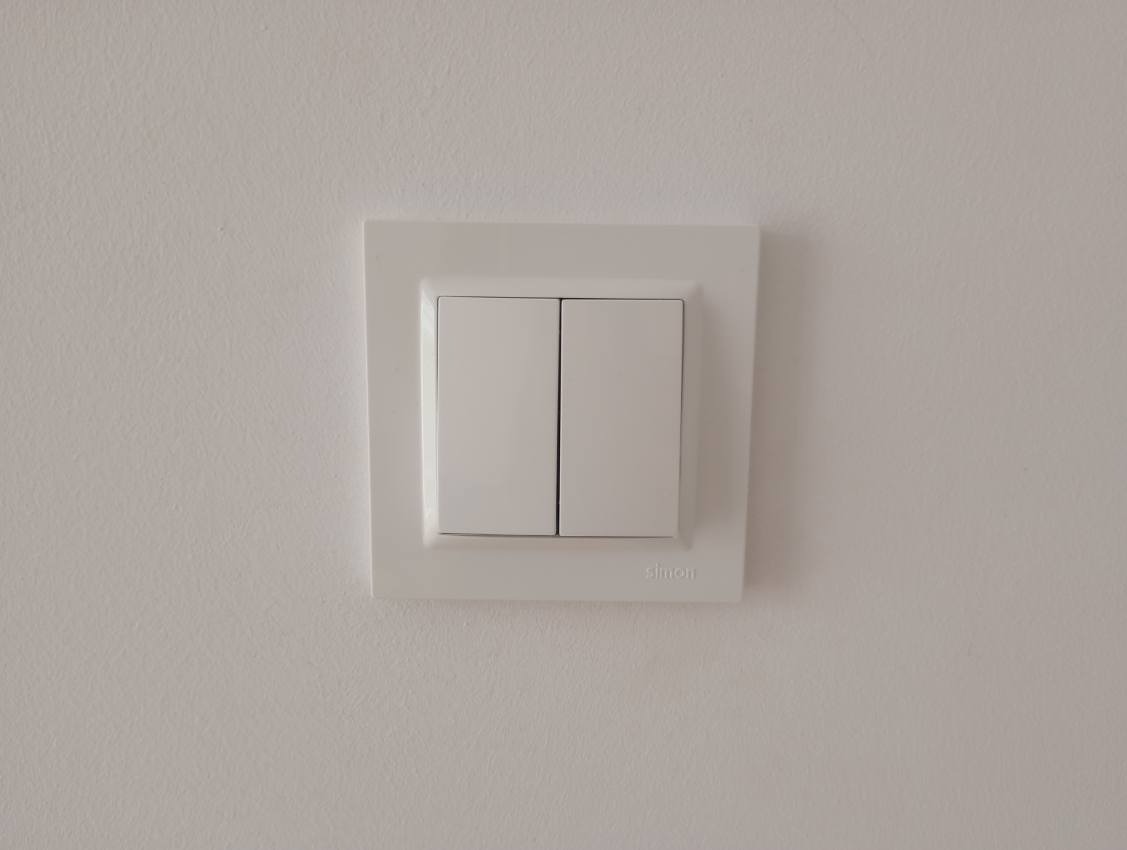 Biały, podwójny włącznik światła marki Simon, zamontowany na jasnej ścianie. Minimalistyczny design w neutralnym wnętrzu.