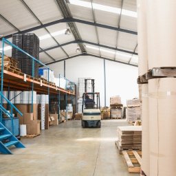 Kartony123 - Wnętrze magazynu w Kórniku z wózkiem widłowym transportującym towary, widoczne rolki papieru i regały z zapasami.