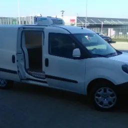 Biały samochód dostawczy z otwartymi drzwiami bocznymi i agregatem chłodniczym na dachu, zaparkowany na brukowanym placu przed budynkiem.