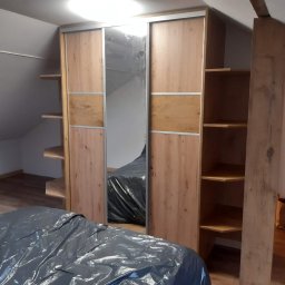 Adrian Zalewski - Montaż szafy przesuwnej z lustrem w skosie poddasza, widoczne półki boczne i zabezpieczone folią meble, surowy drewniany słup podtrzymujący.
