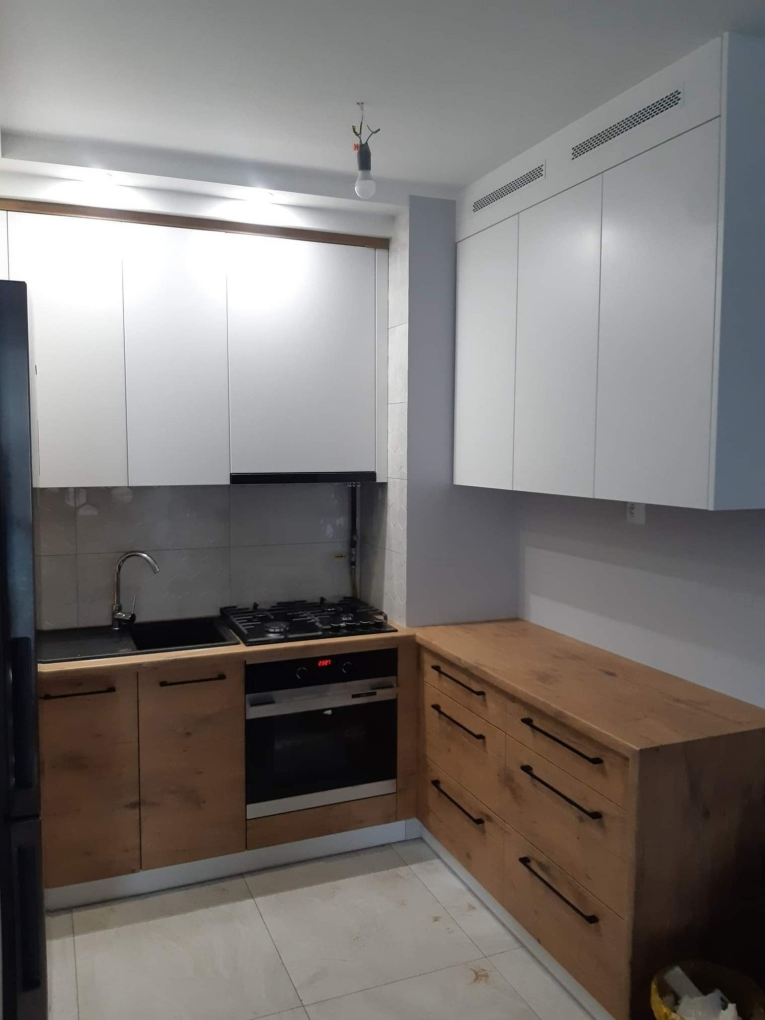 Nowoczesna kuchnia z białymi szafkami górnymi i drewnianymi szafkami dolnymi z czarnymi uchwytami, widoczny piekarnik i płyta gazowa, minimalistyczny styl, niedokończone oświetlenie sufitowe.