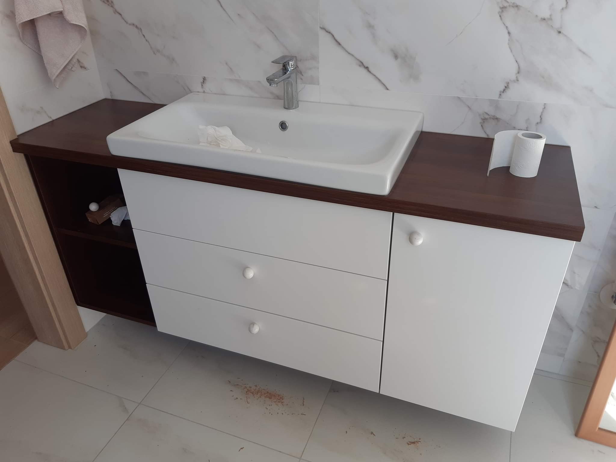 Biała umywalka nablatowa z trzema szufladami i brązowym blatem, zamontowana w łazience z marmurowymi płytkami na ścianie. Widoczny dozownik mydła i rolka papieru toaletowego.