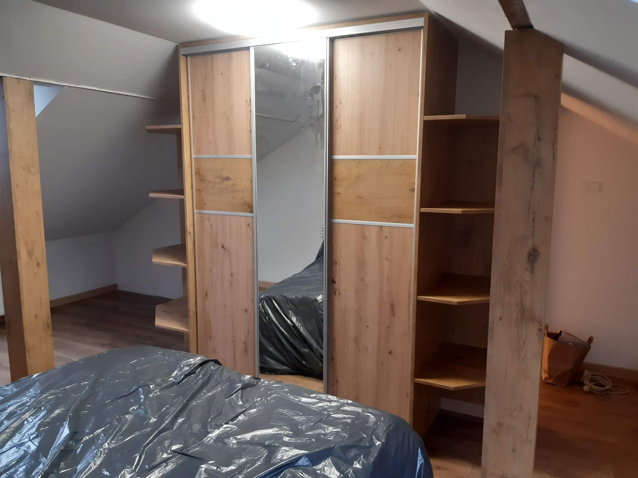 Montaż szafy przesuwnej z lustrem w skosie poddasza, widoczne półki boczne i zabezpieczone folią meble, surowy drewniany słup podtrzymujący.