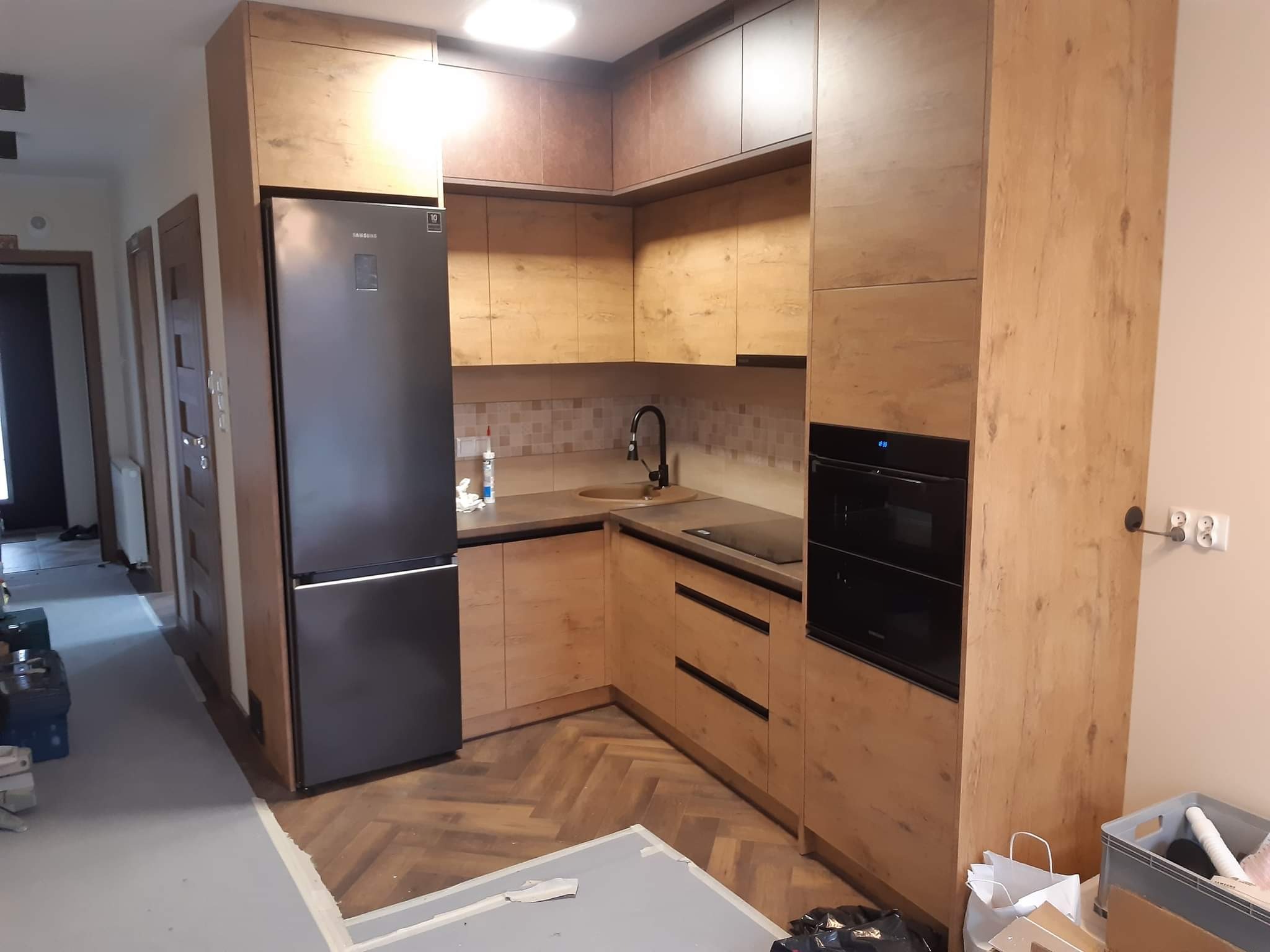 Kuchnia na wymiar z drewnianymi frontami, czarnym sprzętem AGD i parkietem w jodełkę, widoczne ślady prac wykończeniowych na podłodze.