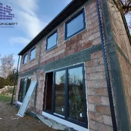 Dom w trakcie budowy z zamontowanymi czarnymi oknami, widoczne cegły i niezakończone prace wykończeniowe, na trawniku oparta szyba okienna, logo firmy w lewym górnym rogu.