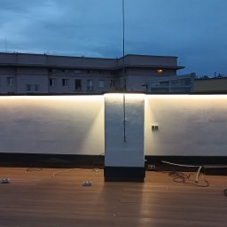 ELWAS ŁUKASZ WĄSIK - Oświetlenie LED zamontowane na ścianie na dachu budynku, widoczne kable i elementy montażowe na drewnianym podłożu, w tle zabudowa miejska pod wieczornym niebem.