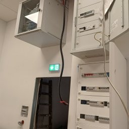 ELWAS ŁUKASZ WĄSIK - Wnętrze w trakcie prac elektrycznych: rozdzielnice elektryczne, szafa serwerowa pod sufitem, drabina oparta o ścianę, widoczny kabel zasilający.