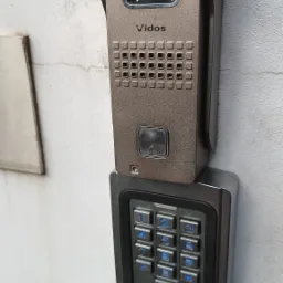 Zewnętrzny panel wideodomofonu Vidos z kamerą i klawiaturą numeryczną zamontowany na białej ścianie. Klawiatura ma podświetlane na niebiesko cyfry.
