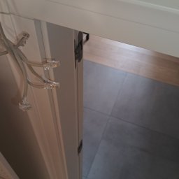 ELWAS ŁUKASZ WĄSIK - Kable sieciowe Ethernet z wtyczkami RJ45 wychodzące z szafy, widok z góry na podłogę z płytek i fragment mebli.