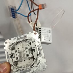 ELWAS ŁUKASZ WĄSIK - Instalacja elektryczna: widok na puszkę instalacyjną z wystającymi przewodami połączonymi złączkami WAGO, w dłoni metalowa ramka gniazdka z widocznymi elementami mocującymi.