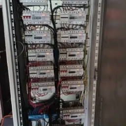 Wnętrze szafy elektrycznej z wieloma licznikami energii, widoczne przewody i zabezpieczenia, oświetlenie LED na boku szafy.