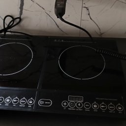 ELWAS ŁUKASZ WĄSIK - Czarna, dwupalnikowa płyta indukcyjna marki Esperanza na blacie kuchennym, z widocznym panelem sterowania i podłączonym zasilaniem. Obok leży miernik elektryczny z pomiarami. Tło stanowi marmurowa...