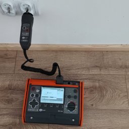 ELWAS ŁUKASZ WĄSIK - Miernik Sonel MPE-530 z podłączonym adapterem w gniazdku elektrycznym, leżący na podłodze z paneli.