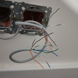 ELWAS ŁUKASZ WĄSIK - Szary kabel sieciowy z rozplątanymi kolorowymi żyłami wystającymi ze ściany, widoczne metalowe puszki montażowe gniazdek.