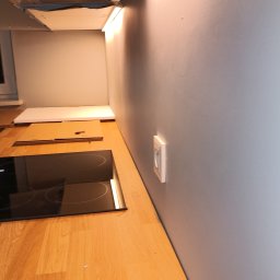 ELWAS ŁUKASZ WĄSIK - Częściowo zamontowana płyta indukcyjna na drewnianym blacie kuchennym, widoczny okap i gniazdko elektryczne na szarej ścianie, oświetlenie podszafkowe włączone.