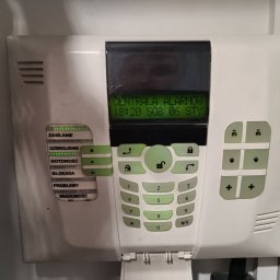 ELWAS ŁUKASZ WĄSIK - Biała centrala alarmowa z zielonym wyświetlaczem i przyciskami, widoczne kontrolki LED stanu systemu, zamontowana na szarej ścianie.