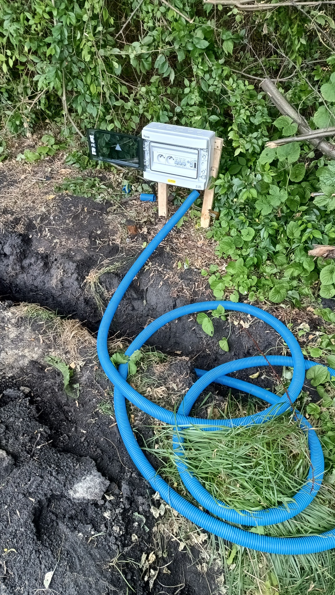 Szara skrzynka elektryczna zamontowana na drewnianych słupkach obok wykopu, z niebieską rurą karbowaną prowadzącą do ziemi, otoczona zielenią.