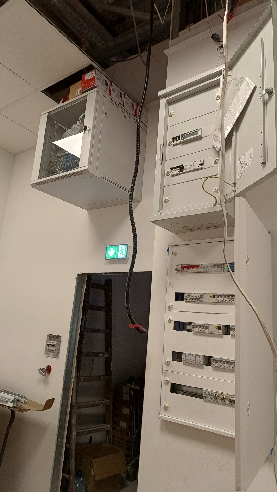 Wnętrze w trakcie prac elektrycznych: rozdzielnice elektryczne, szafa serwerowa pod sufitem, drabina oparta o ścianę, widoczny kabel zasilający.
