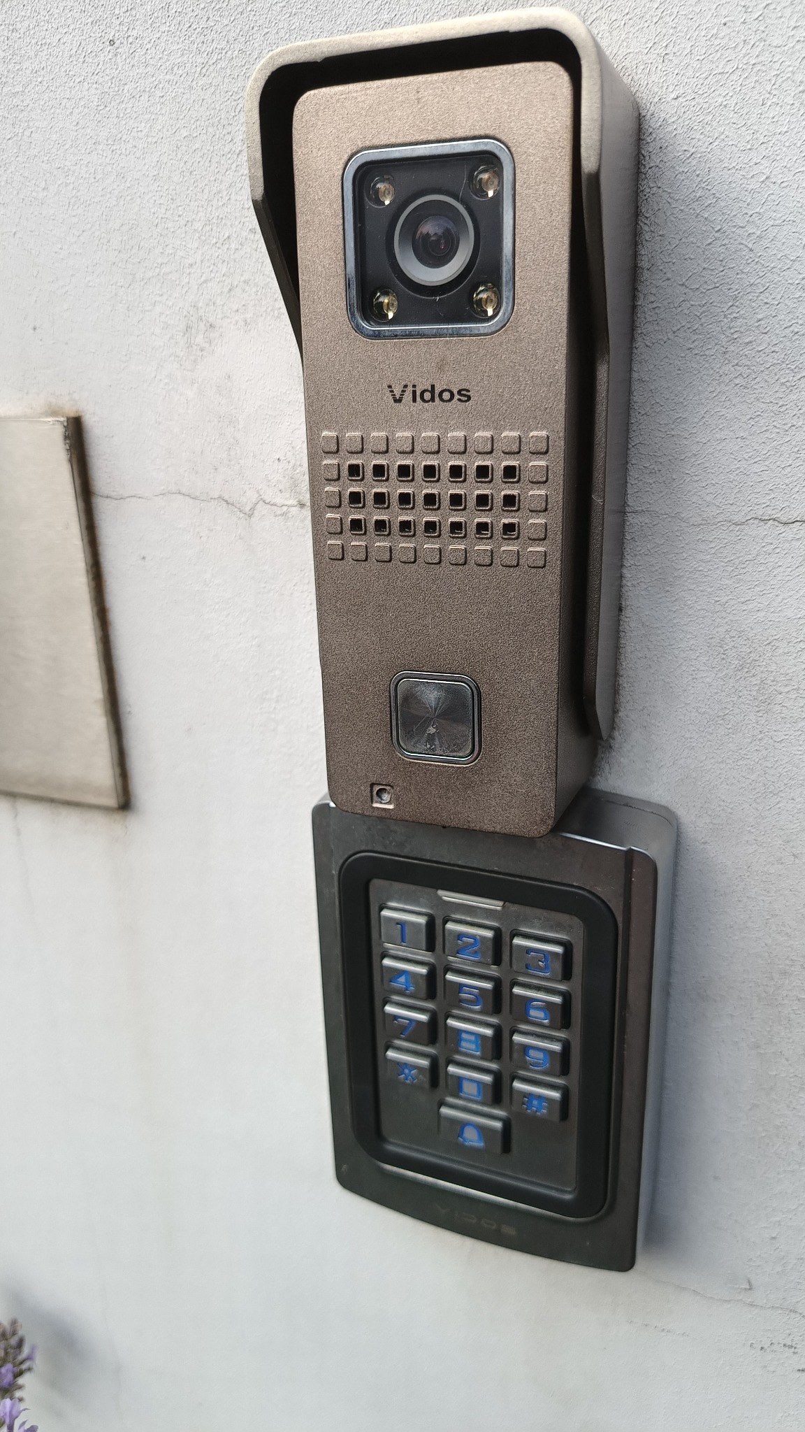 Zewnętrzny panel wideodomofonu Vidos z kamerą i klawiaturą numeryczną zamontowany na białej ścianie. Klawiatura ma podświetlane na niebiesko cyfry.