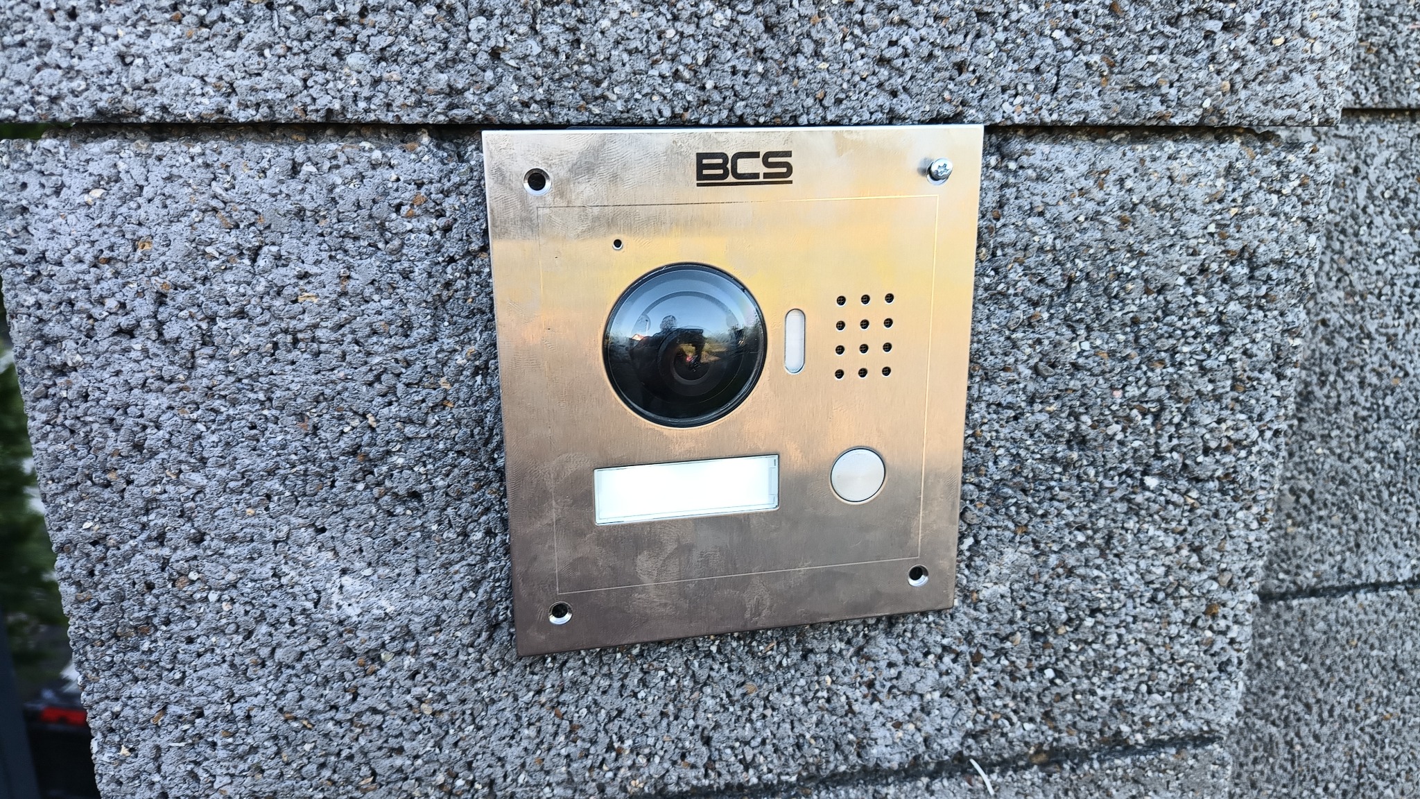Panel domofonowy BCS z kamerą, zamontowany na szarej, chropowatej ścianie z betonu komórkowego, widoczne ślady montażu.