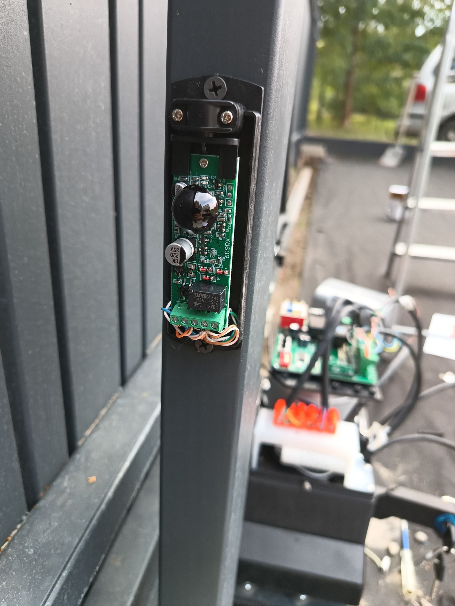 Czujnik zbliżeniowy bramy automatycznej z widoczną elektroniką i okablowaniem, zamontowany na słupku bramy w Warszawie, z elementami mechanizmu napędu w tle.