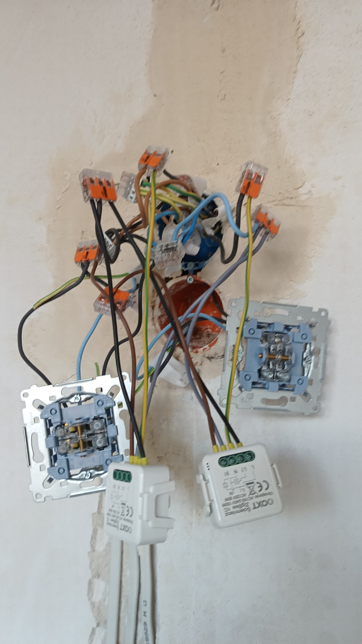 Rozgałęziona instalacja elektryczna z przewodami w różnych kolorach, kostkami WAGO i dwoma białymi modułami sterującymi DOXT ZigBee 1C, widoczne puszki instalacyjne w ścianie w trakcie prac.