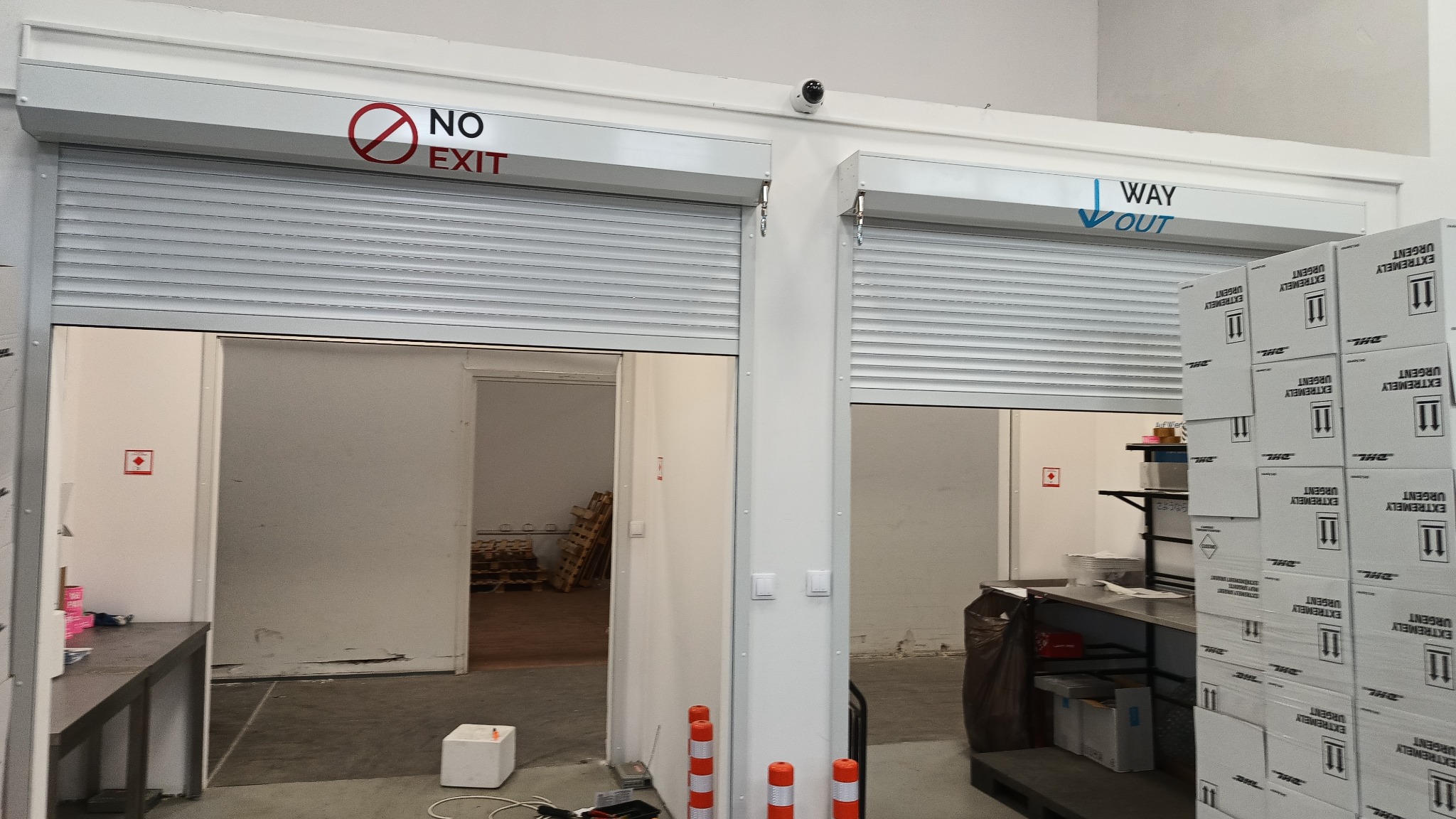 Dwa wejścia do magazynu z roletami, jedno z napisem 'NO EXIT' i przekreślonym kołem, drugie z napisem 'WAY OUT', nad nimi kamera przemysłowa, w tle widoczne palety i kartony z napisem 'EXTREMELY...
