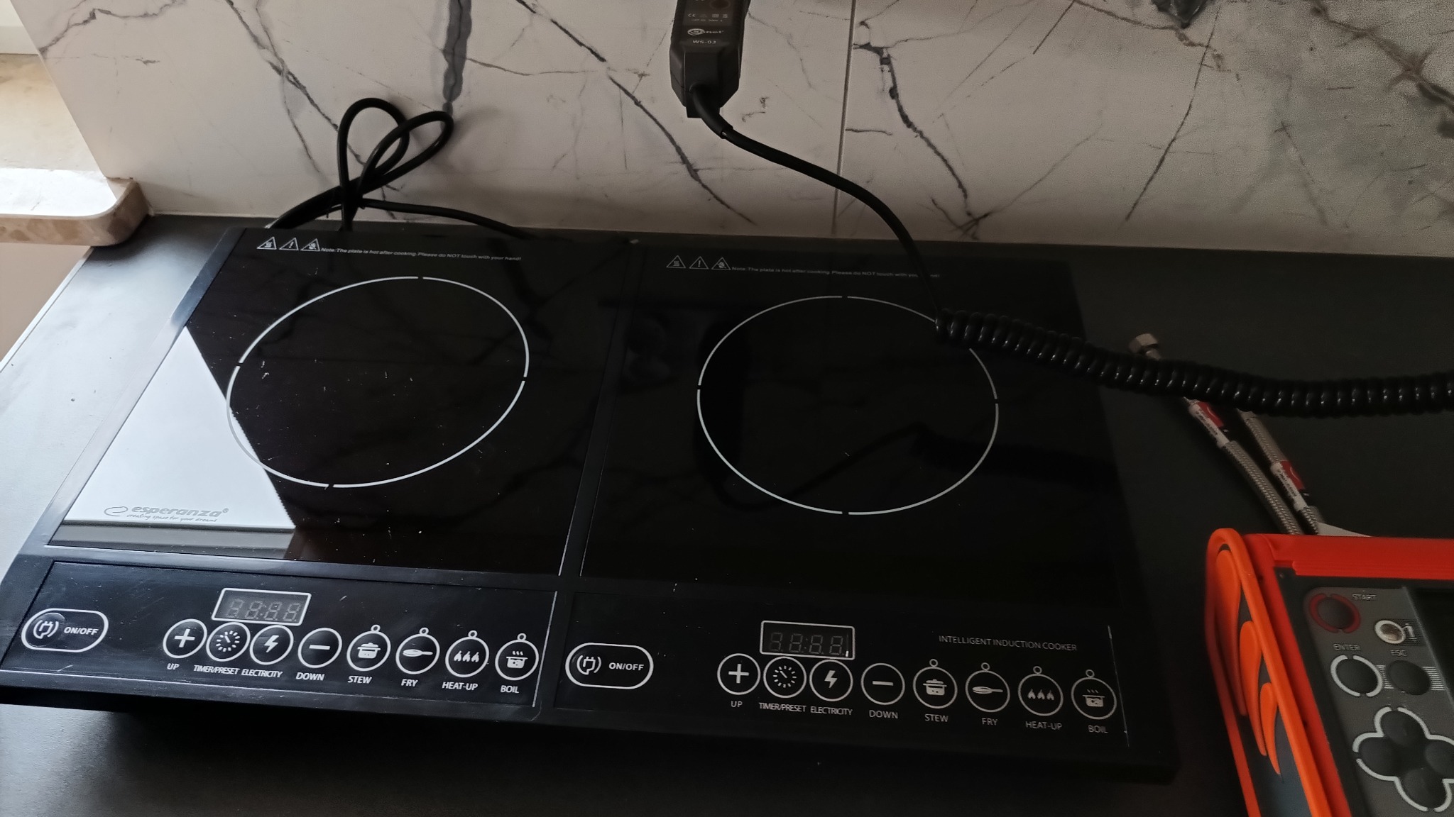 Czarna, dwupalnikowa płyta indukcyjna marki Esperanza na blacie kuchennym, z widocznym panelem sterowania i podłączonym zasilaniem. Obok leży miernik elektryczny z pomiarami. Tło stanowi marmurowa...